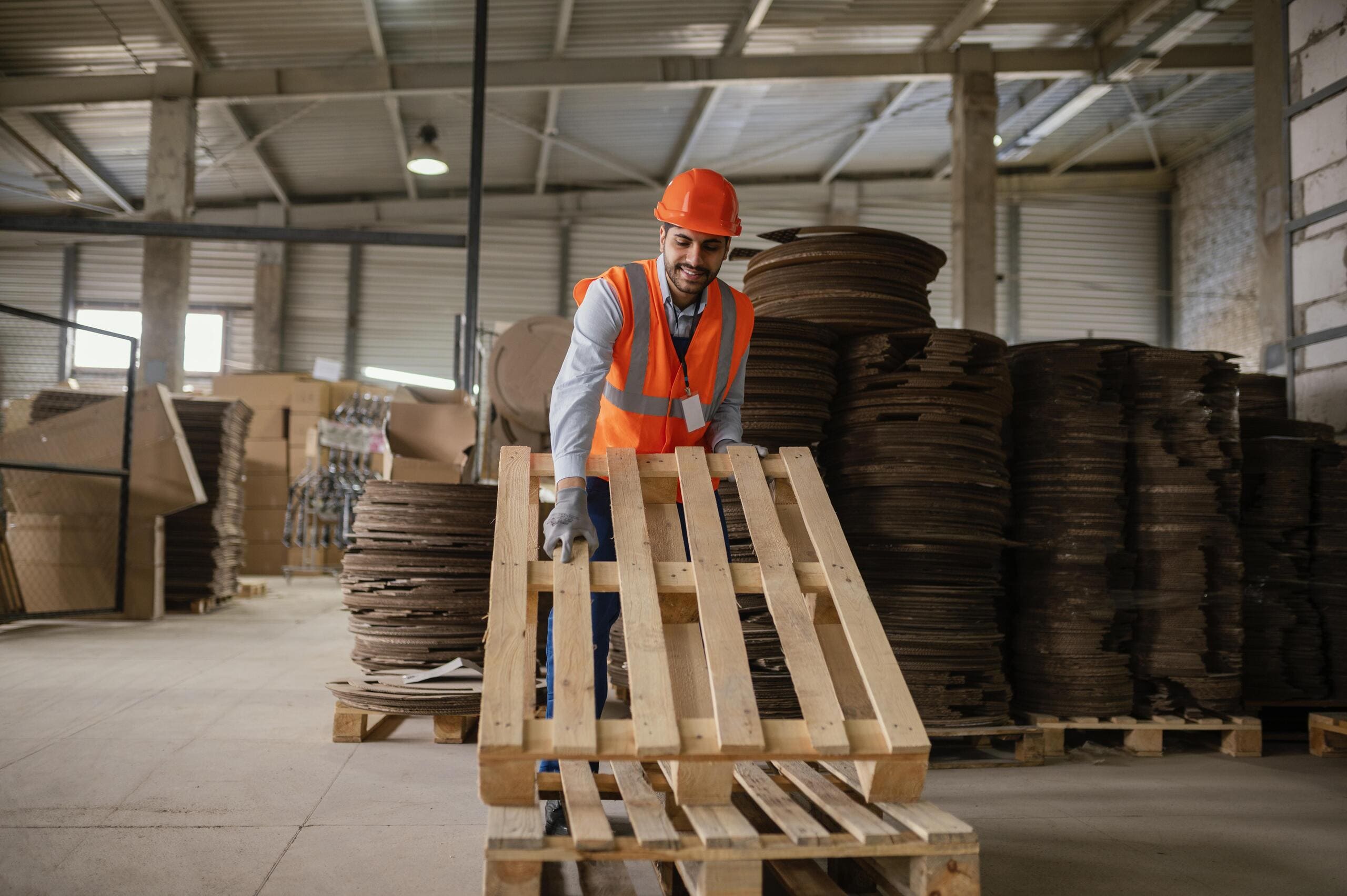 Trabalhador com colete e capacete manuseando pallet novos ou usados de madeira em galpão logístico.